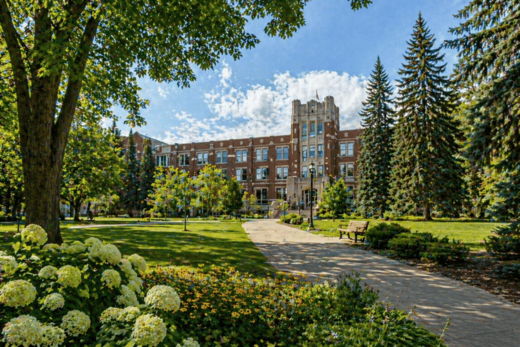 university campus with red brick building, green lawn, trees, flowers, and pathway under clear blue sky